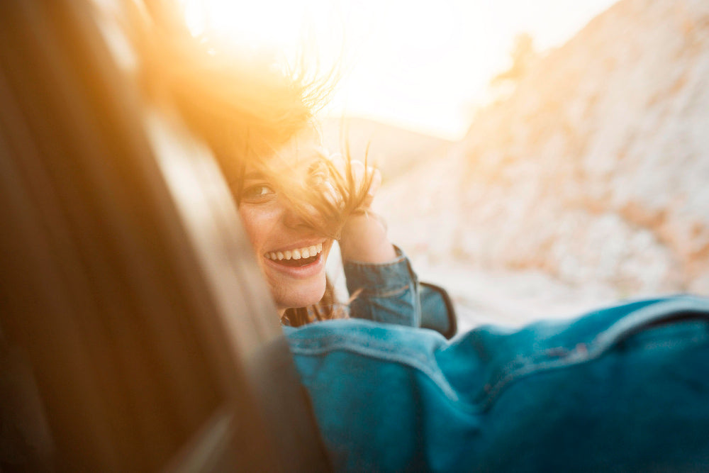 A smiling woman in a blue denim jacket and jeans lies on a snowy hillside outdoors, with bright warm sunlight and lens flare creating a dreamy, backlit scene.